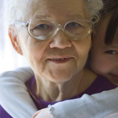 An older woman with white hair and glasses looks at the viewer. A girl of around 5-6 years old has her arms around her neck affectionately. 